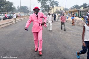 an image from Tamagni's book that displays a sapeur donning a bright pink suit, pink hat, and pink shoes with a cigar in his mouth struts on a dusty concrete road with cars passing on his left and regularly dressed people behind him. 