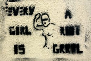 Black graffiti on a white wall that reads "Every girl is a Riot Grrrl" with an outline of a naked woman holding up one of her arms.