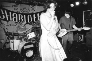 A black and white image of a woman in a dress and glasses singing into a mircophone. Another band member in the background is playing the electric guitar while another woman plays the drums.
