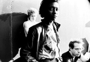This is a black and white picture of Karla Mad Dog standing in the center, facing the camera, with her hands in her pockets. Behind her, slightly to the left is another band member with their back against the wall, folding their arms. In the bottom right corner of the photo, is the head of the third members, who appears to be sitting down. His face cannot be fully seen.