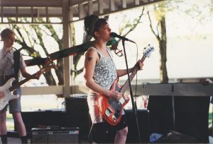 An image of Kathleen Hanna playing electric guitar and singing into a microphone on stage. Another member of Bikini Kill also plays electric guitar in the background.