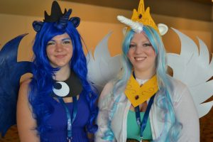 Image of two people dressed up as My Little Pony characters. The two people have colorful wigs on, wings, and horns.