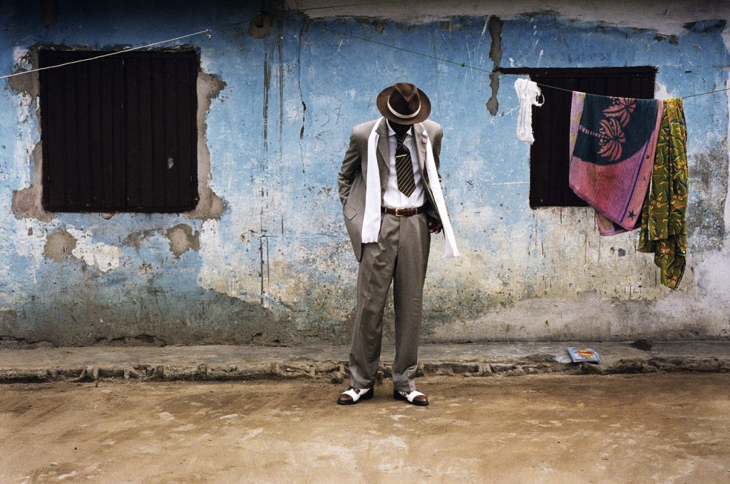 A sapeur wearing a three-colored coordinated suit of ash brown, white, and dark brown. His face is hidden by a wide-brimmed brown hat as he stares towards his designer leather shoes. His figure and attire is in stark contrast to the faded wall of a dilapidated structure behind him. The dirt road he stands on is dusty and there is a makeshift laundry string hanging above his head with towels. 