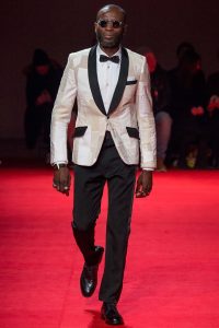 one of Watanabe's models walking down the red carpet with a more "traditional" white dress shirt and black pants and shoes. The reminiscent quality of sapeur fashion is found in the nuances of the jacket, adherence to the "three-color" rule, and general atmosphere of the model. He wears circular sunglasses, a black bowtie, and a white gold metallic jacket with black lapels, pocket covers, and handkerchief pocket. 