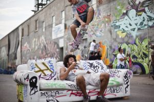 A skater does a trick over a couch while another man sits on the couch. The couch and the walls behind it are covered in graffiti.