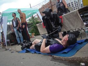man wearing leather lays down on the floor holding the leash of a man dressed in a full puppy play leather outfit. Others stand in the background at this outside street festival.