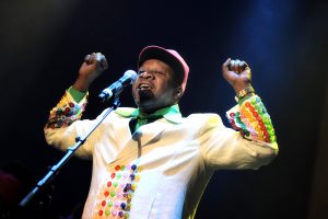 Papa Wemba performs in front of a microphone, his hands theatrically popped up at his sides. He is wearing a cap, pale yellow suit jacket with multi-colored buttons all over the cuffs and the front of his jacket. 