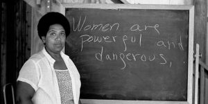 A black and white photograph of Audre Lorde standing in front of a blackboard that has "Women are powerful and dangerous" handwritten across it in chalk.