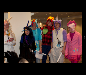 An image of six bronies dressed as characters from the show. Four of the fans are male. Each of the fans is dressed in a different costume involving bright colors, pony ears, and a unicorn horn. 