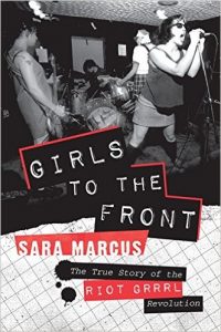 An image of the cover of "Girls to Front: The True Story of the Riot Grrrl Revolution" featuring a black and white photograph of a Riot Grrrl band performing on stage.