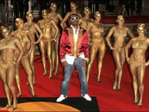 A photo of a male hip hop artist surrounded by scantily clad women who are covered in gold glitter as if they're trophies
