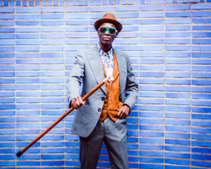 man with blue sunglasses, brown hat, gray pinstripe suit, colorful shirt and brown vest