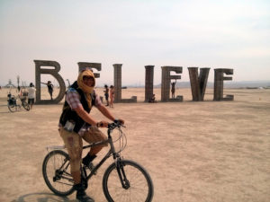 A 'Burner' cycling around the desert site filled with large scale art installations; this image shows a n installation that spells out the word 'believe' in large letters