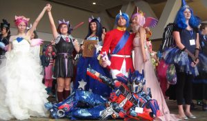 Photoshoot at BronyCon in which a group of people are dressed as characters from the show. For example, one woman is dressed in all pink with a horn and a man is dressed in a uniform and a horn 