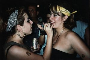 One woman stands in front of another adjusting her makeup.