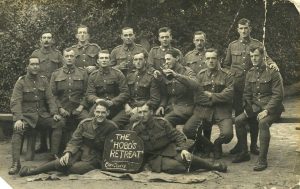 A large group photo of many young men all wearing the same uniform of dark pants with button downs. The men sitting in the front and middle hold a sign saying, “The Hobo Retreat”.