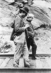 Black and white photo of two older men walk down train tracks in front of stone and they are dark gray wearing jackets. One wears a black fedora and is carrying a rucksack and the other wears a white conductor hat.