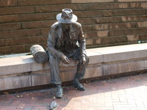 A bronze statue of a man sitting wearing an overcoat and wide brimmed hat. There is a bronze rolled blanket next to him and he is staring at a tiny bronze duck by his left foot.