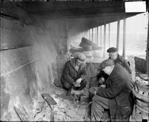 Three older men sit in a circle in a broken down building. They are all wearing black jackets and gray fedoras.