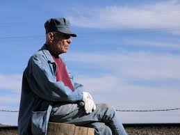 An older man sits cross-legged on fencepost in front of a blue sky with clouds. He is wearing a red T-shirt with a denim shirt over it and jeans. He is also wearing white gloves and a blue conductor hat.