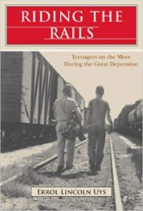 A black and white photo of two young men walking down railroad tracks with their back to the camera. The man on the left holds a plaid blanket. The one on the right holds a small bundle of clothes. There are trains on the tracks on either side of the men. 