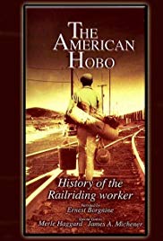 A man walks away from the camera down traintracks. He has a rolled blanket on his back and is holding a bucket in his left hand and a bag in his right. There is a golden light filter.