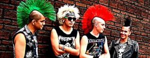 Four young men with brightly colored hair and black clothing stand in front of a brick wall.