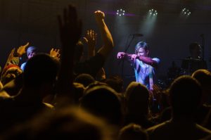 View of Alternative Rock performance from the perspective of an audience member, band member points toward crowd from under the spotlight
