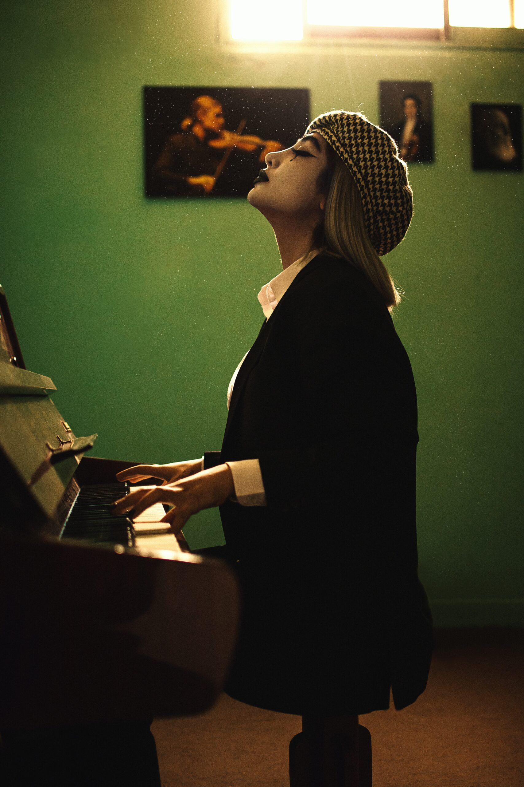 A woman in a black blazer plays the piano with a pensive expression. There are portraits of musicians in the background.